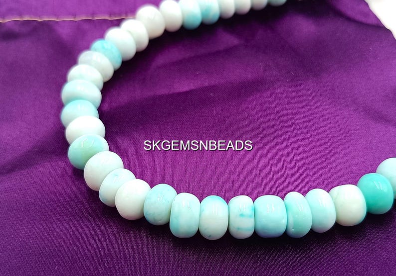 May include: A necklace of round, light blue and white beads. The beads are strung together, creating a curved shape against a purple background. The text "SKGEMSNEADS" is visible.