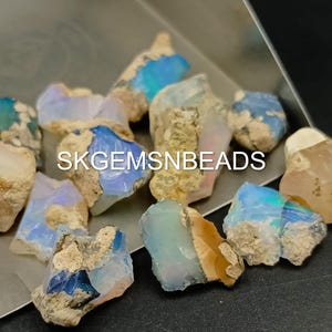 Puede incluir: Una colecci&oacute;n de piedras preciosas de &oacute;palo en bruto en varios tonos de azul, blanco y marr&oacute;n. Los &oacute;palos tienen una forma irregular y una apariencia natural y en bruto. SKGEMSNBEADS est&aacute; impreso en la imagen.