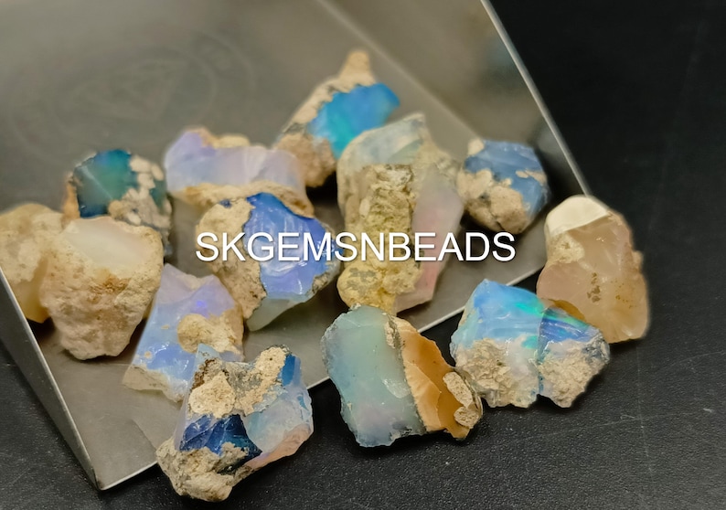Puede incluir: Una colecci&oacute;n de piedras preciosas de &oacute;palo en bruto en varios tonos de azul, blanco y marr&oacute;n. Los &oacute;palos tienen una forma irregular y una apariencia natural y en bruto. SKGEMSNBEADS est&aacute; impreso en la imagen.