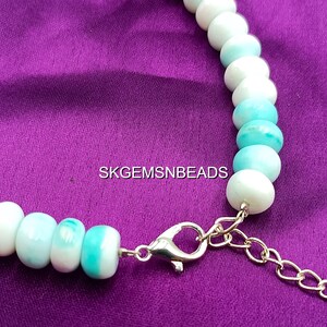 May include: A necklace featuring round, light blue and white beads. The beads are strung together with a silver clasp and a gold-colored chain extender. The necklace is displayed on a purple fabric background. The text "SKGEMSNBEADS" is visible.