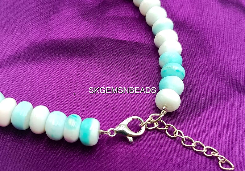 May include: A necklace featuring round, light blue and white beads. The beads are strung together with a silver clasp and a gold-colored chain extender. The necklace is displayed on a purple fabric background. The text "SKGEMSNBEADS" is visible.