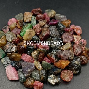 K&ouml;nnte beinhalten: Eine Sammlung von bunten, rohen Edelsteinen, darunter rosa, rot, gr&uuml;n und schwarz. Die Edelsteine sind auf einer dunklen Oberfl&auml;che verstreut. SKGEMSNBEADS