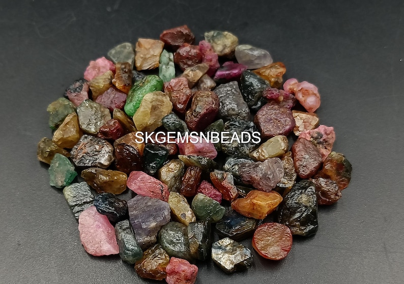 K&ouml;nnte beinhalten: Eine Sammlung von bunten, rohen Edelsteinen, darunter rosa, rot, gr&uuml;n und schwarz. Die Edelsteine sind auf einer dunklen Oberfl&auml;che verstreut. SKGEMSNBEADS