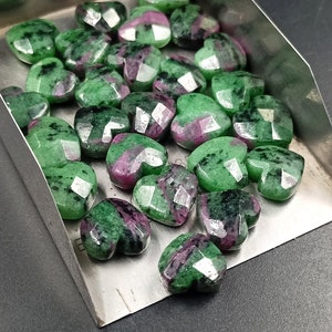 May include: A collection of green and pink heart-shaped gemstones. The stones have a faceted finish and are arranged in a metal tray.