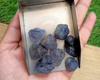 May include: A collection of rough, dark blue-purple natural stones displayed in a small, rectangular metal tray. The tray is held in a hand, with a green background. The tray has the words "NATURAL COLOR STONE" printed on it.