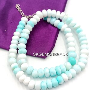 May include: A necklace composed of white and light blue beads. The beads are strung together with a silver-colored chain and are arranged in a double strand. The necklace is displayed on a purple fabric.