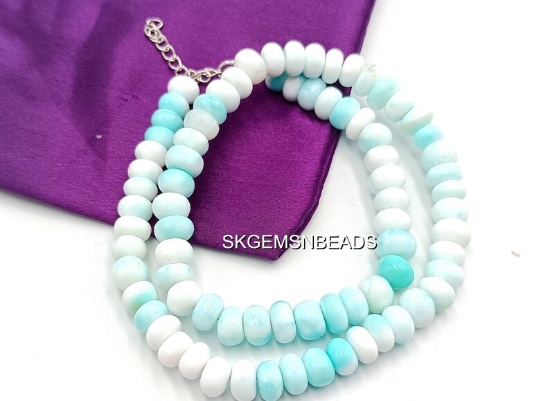 May include: A necklace composed of white and light blue beads. The beads are strung together with a silver-colored chain and are arranged in a double strand. The necklace is displayed on a purple fabric.