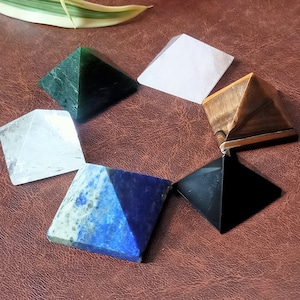 Könnte beinhalten: Eine Sammlung von sieben Edelsteinpyramiden in verschiedenen Farben, darunter Grün, Weiß, Braun, Schwarz, Klar und Blau. Die Pyramiden sind kreisförmig auf einer braunen Oberfläche angeordnet, mit einem grünen und weißen Blatt im Hintergrund.