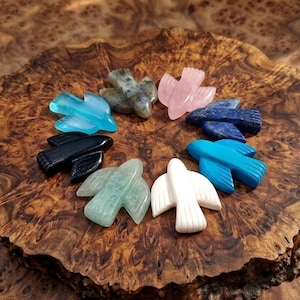 May include: A collection of carved stone bird figurines in various colors, including blue, pink, black, and white, arranged in a circle on a wooden surface. The birds are detailed and appear to be made of different types of stone.