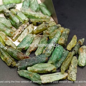 May include: A close-up of a pile of green and brown gemstone chips. The chips are long and thin, and they have a rough, natural texture. The chips are scattered on a dark surface. The text "SKGEMSNBEADS" is visible in the image.