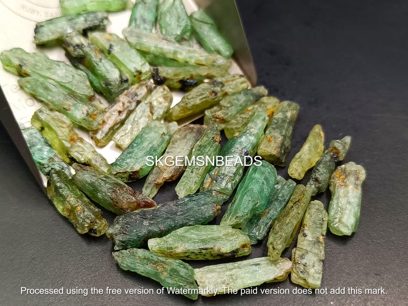 May include: A close-up of a pile of green and brown gemstone chips. The chips are long and thin, and they have a rough, natural texture. The chips are scattered on a dark surface. The text "SKGEMSNBEADS" is visible in the image.