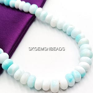 May include: A necklace of round, polished beads in shades of white and light blue. The beads are strung together, creating a curved shape. The necklace is displayed on a purple satin fabric. The text "SKGEMSNBEADS" is visible.