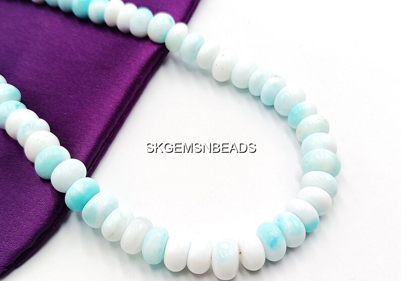 May include: A necklace of round, polished beads in shades of white and light blue. The beads are strung together, creating a curved shape. The necklace is displayed on a purple satin fabric. The text "SKGEMSNBEADS" is visible.