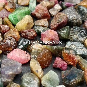 K&ouml;nnte beinhalten: Eine Sammlung von rohen, ungeschliffenen Edelsteinen in verschiedenen Farben, darunter Gr&uuml;n, Blau, Rosa und Rot. Die Edelsteine sind auf einer wei&szlig;en Oberfl&auml;che verstreut. SKGEMSNBEADS ist auf dem Bild gedruckt.