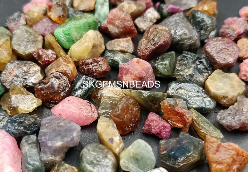 K&ouml;nnte beinhalten: Eine Sammlung von rohen, ungeschliffenen Edelsteinen in verschiedenen Farben, darunter Gr&uuml;n, Blau, Rosa und Rot. Die Edelsteine sind auf einer wei&szlig;en Oberfl&auml;che verstreut. SKGEMSNBEADS ist auf dem Bild gedruckt.