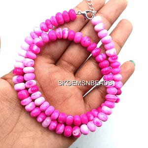May include: A vibrant pink and white beaded necklace with a silver clasp. The necklace is composed of small, oval-shaped beads in varying shades of pink and white. The text "SKGEMSNBEADS" is visible in the background.