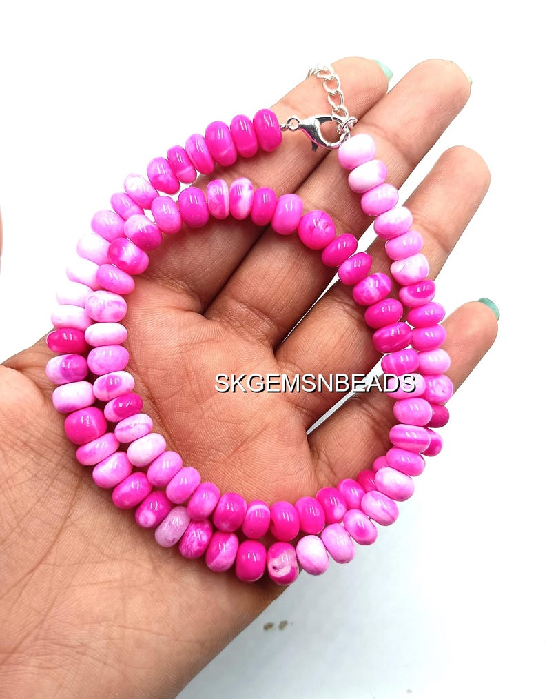 May include: A vibrant pink and white beaded necklace with a silver clasp. The necklace is composed of small, oval-shaped beads in varying shades of pink and white. The text "SKGEMSNBEADS" is visible in the background.