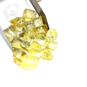 K&ouml;nnte beinhalten: Ein Haufen gelber Citrin-Edelsteine, einige mit einem leichten Orangeton, fallen aus einer silbernen Schaufel. Die Schaufel tr&auml;gt ein Etikett mit der Aufschrift "Natural Color Stone".