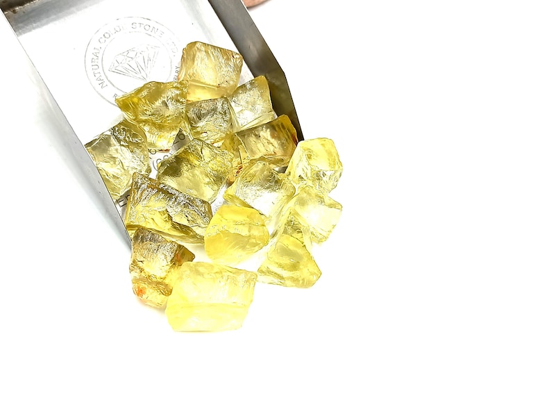 K&ouml;nnte beinhalten: Ein Haufen gelber Citrin-Edelsteine, einige mit einem leichten Orangeton, fallen aus einer silbernen Schaufel. Die Schaufel tr&auml;gt ein Etikett mit der Aufschrift "Natural Color Stone".