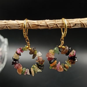 Puede incluir: Aretes de aro dorados con un círculo de piedras preciosas multicolores. Las piedras preciosas son en tonos de rosa, verde, negro y marrón.