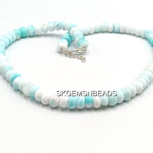 May include: A necklace composed of round, polished beads in shades of white and light blue. The beads are strung together, forming a circular shape with a silver clasp. The necklace is displayed on a white surface.