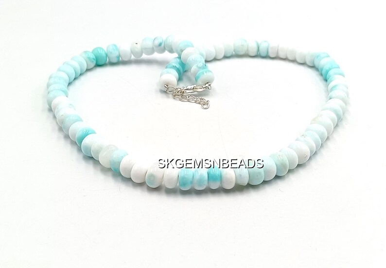 May include: A necklace composed of round, polished beads in shades of white and light blue. The beads are strung together, forming a circular shape with a silver clasp. The necklace is displayed on a white surface.