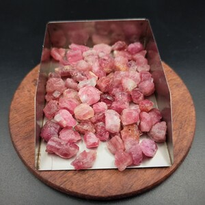 May include: A close-up of a pile of pink and red rough gemstones in a metal tray. The gemstones are various sizes and shapes.