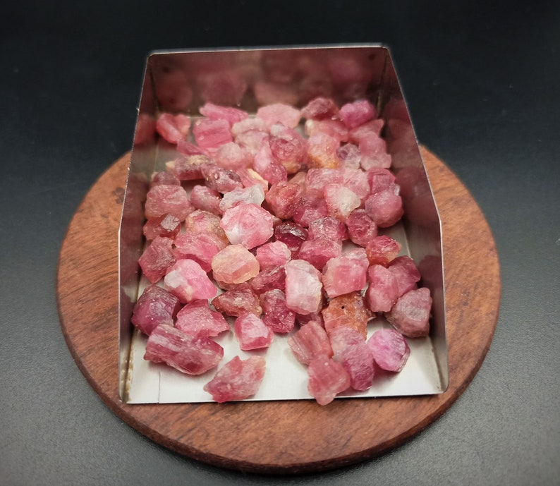 May include: A close-up of a pile of pink and red rough gemstones in a metal tray. The gemstones are various sizes and shapes.