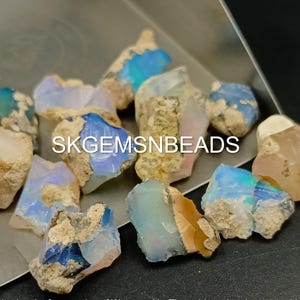 Puede incluir: Una colecci&oacute;n de piedras preciosas de &oacute;palo natural en bruto en varios tonos de azul, blanco y marr&oacute;n. Las piedras tienen una forma irregular y una textura rugosa y sin pulir. El texto "SKGEMSNBEADS" es visible en la imagen.