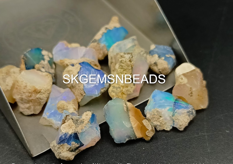 Puede incluir: Una colecci&oacute;n de piedras preciosas de &oacute;palo natural en bruto en varios tonos de azul, blanco y marr&oacute;n. Las piedras tienen una forma irregular y una textura rugosa y sin pulir. El texto "SKGEMSNBEADS" es visible en la imagen.