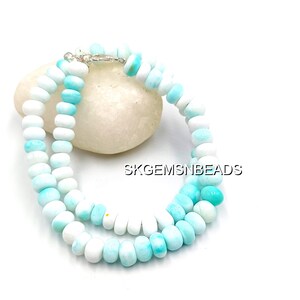 May include: A necklace made of white and light blue beads. The beads are round and strung together with a silver clasp. The necklace is resting on a white stone. The text "SKGEMSNBEADS" is visible.