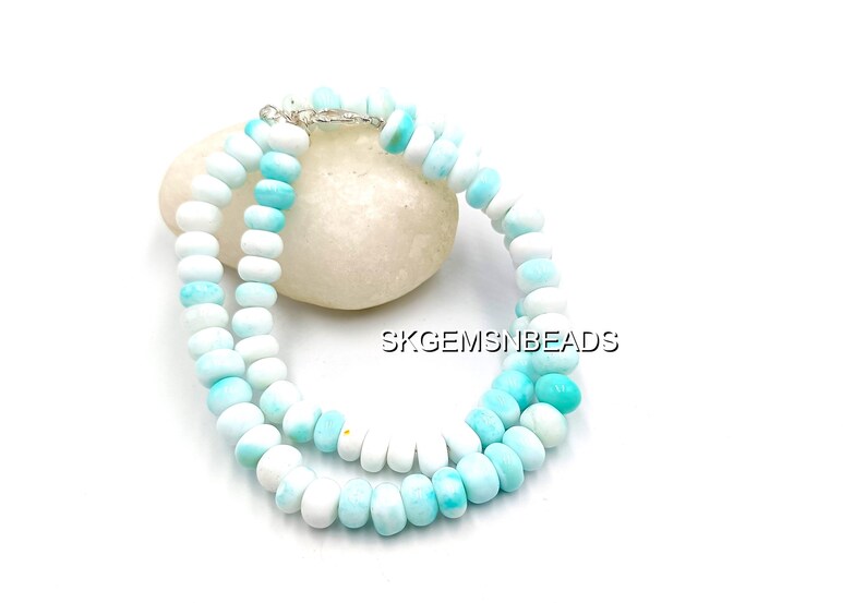 May include: A necklace made of white and light blue beads. The beads are round and strung together with a silver clasp. The necklace is resting on a white stone. The text "SKGEMSNBEADS" is visible.