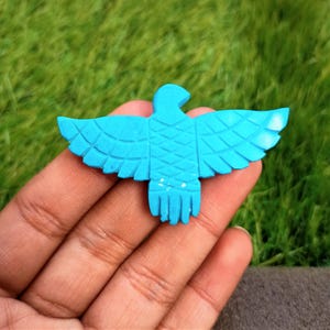 May include: A turquoise eagle figurine with outstretched wings. The bird is carved with detailed feather patterns and a textured body. The figurine is held in a hand against a backdrop of green grass.