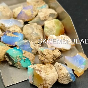 Puede incluir: Una colecci&oacute;n de piedras preciosas de &oacute;palo natural en bruto en varios tonos de azul, verde y blanco. Los &oacute;palos est&aacute;n colocados sobre un fondo plateado. SKGEMSNBEADS est&aacute; impreso en el fondo.