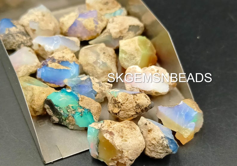 Puede incluir: Una colecci&oacute;n de piedras preciosas de &oacute;palo natural en bruto en varios tonos de azul, verde y blanco. Los &oacute;palos est&aacute;n colocados sobre un fondo plateado. SKGEMSNBEADS est&aacute; impreso en el fondo.