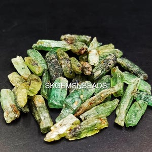 May include: A pile of green and brown rough emerald crystals. The crystals are long and thin, with a pointed end. They are arranged in a random pattern. SKGEMSNBEADS is printed on the image.