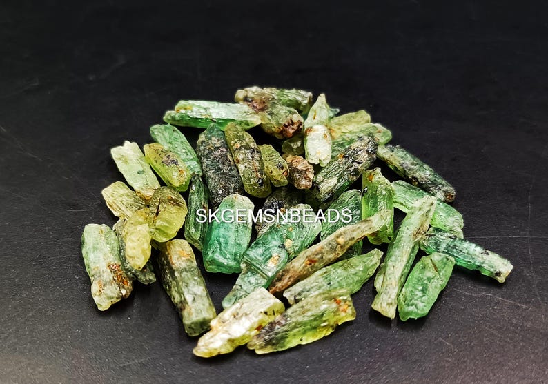 May include: A pile of green and brown rough emerald crystals. The crystals are long and thin, with a pointed end. They are arranged in a random pattern. SKGEMSNBEADS is printed on the image.