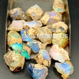 Puede incluir: Una colecci&oacute;n de piedras preciosas de &oacute;palo en bruto en varios tonos de azul, verde, blanco y rosa. Los &oacute;palos est&aacute;n en una bandeja de metal y tienen una apariencia &aacute;spera y natural. SKGEMSNBEADS est&aacute; impreso en la bandeja.
