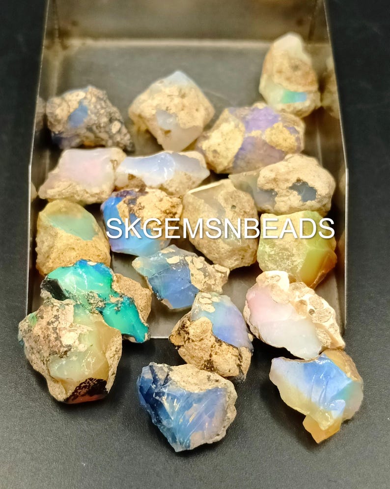 Puede incluir: Una colecci&oacute;n de piedras preciosas de &oacute;palo en bruto en varios tonos de azul, verde, blanco y rosa. Los &oacute;palos est&aacute;n en una bandeja de metal y tienen una apariencia &aacute;spera y natural. SKGEMSNBEADS est&aacute; impreso en la bandeja.