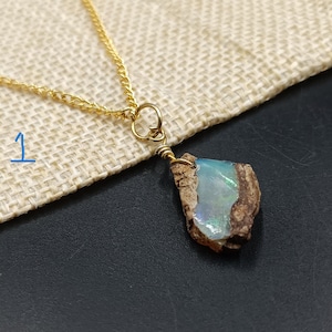 Op de afbeelding: Een goudkleurige ketting met een hanger van ruwe opaal. De opaal heeft een lichtblauwe en iriserende uitstraling, gezet in een draadwikkel. De hanger is met een goudkleurige ring aan de ketting bevestigd.