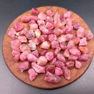 May include: A collection of pink ruby rough stones on a wooden surface. The stones are various sizes and shapes, with some having a more translucent appearance than others.