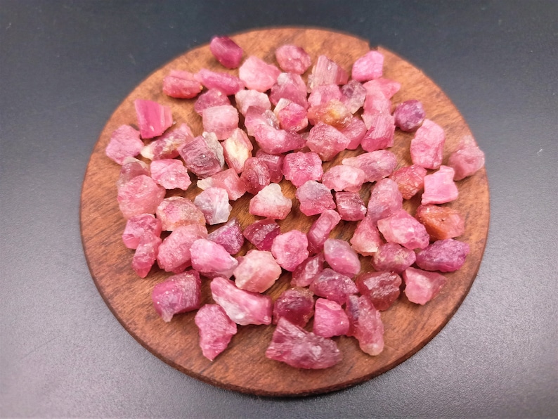 May include: A collection of pink ruby rough stones on a wooden surface. The stones are various sizes and shapes, with some having a more translucent appearance than others.