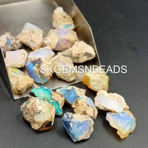Puede incluir: Una colecci&oacute;n de piedras preciosas de &oacute;palo en bruto de varios colores, incluidos azul, verde y blanco. Los &oacute;palos est&aacute;n en una pala de metal y esparcidos sobre una superficie oscura. El texto "SKGEMSNBEADS" es visible en la imagen.