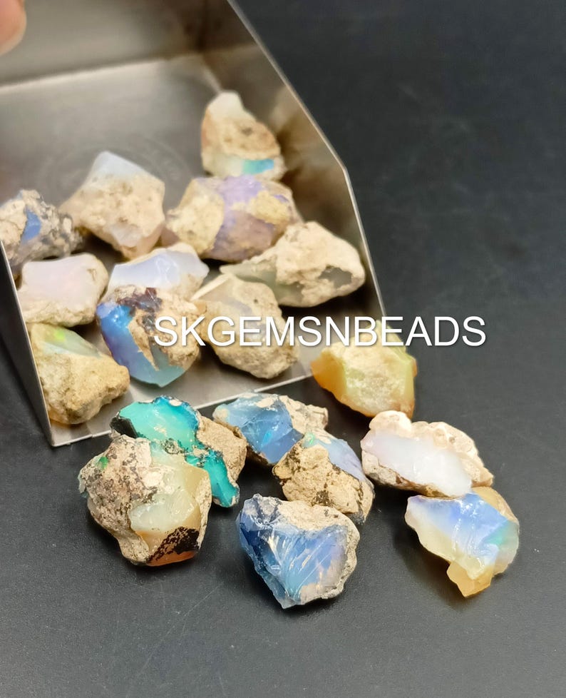 Puede incluir: Una colecci&oacute;n de piedras preciosas de &oacute;palo en bruto de varios colores, incluidos azul, verde y blanco. Los &oacute;palos est&aacute;n en una pala de metal y esparcidos sobre una superficie oscura. El texto "SKGEMSNBEADS" es visible en la imagen.