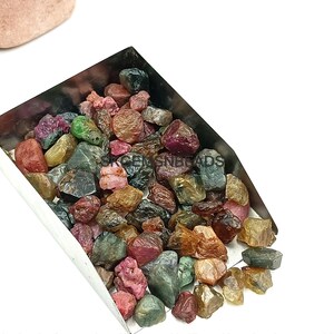 K&ouml;nnte beinhalten: Eine Metallschaufel gef&uuml;llt mit einer Vielzahl von bunten Edelsteinen. Die Edelsteine sind eine Mischung aus Gr&uuml;n, Rot, Rosa und Gelb. Die Edelsteine sind alle roh und unpoliert.