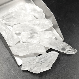 May include: A close-up of a pile of clear quartz crystals in a metal scoop. The crystals are various sizes and shapes, with some having sharp edges.