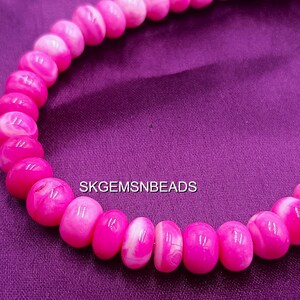 May include: A strand of pink and white beads, likely for jewelry making, against a purple background. The beads are oval-shaped and have a glossy finish. The text "SKGEMSNBEADS" is visible.
