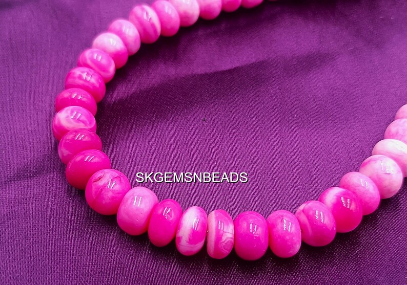 May include: A strand of pink and white beads, likely for jewelry making, against a purple background. The beads are oval-shaped and have a glossy finish. The text "SKGEMSNBEADS" is visible.
