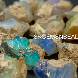 Puede incluir: Una colecci&oacute;n de piedras de &oacute;palo en bruto en varios tonos de azul, verde y blanco. Las piedras se colocan sobre un fondo oscuro. SKGEMSNBEADS est&aacute; impreso en la imagen.