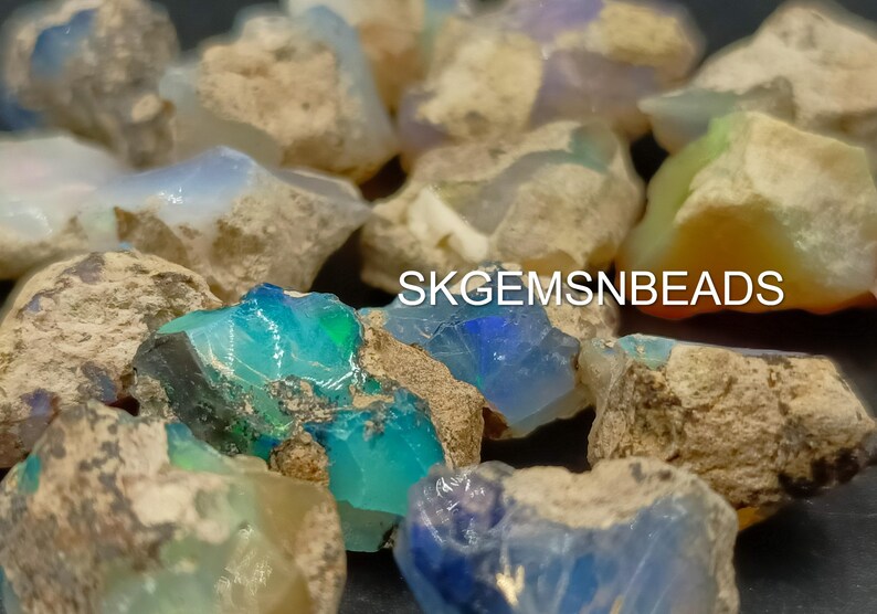 Puede incluir: Una colecci&oacute;n de piedras de &oacute;palo en bruto en varios tonos de azul, verde y blanco. Las piedras se colocan sobre un fondo oscuro. SKGEMSNBEADS est&aacute; impreso en la imagen.